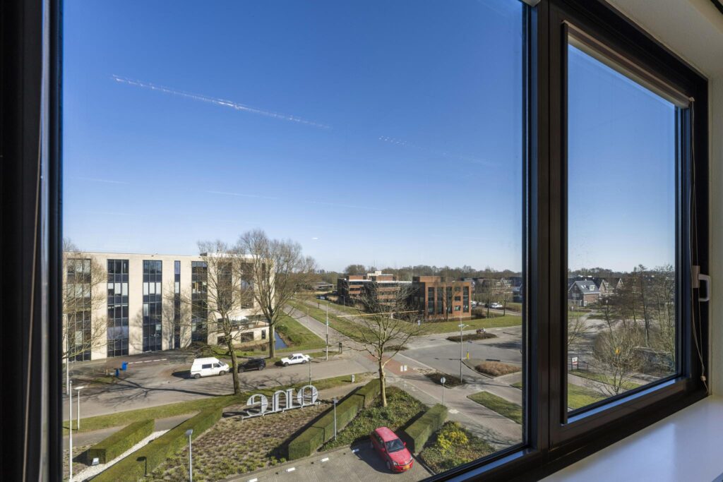 View from a window overlooking Prof. W.H. Keesomlaan with office buildings, parked cars, and a clear blue sky.